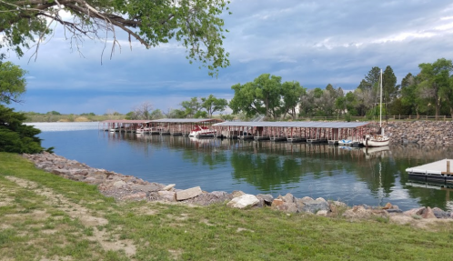 A calm lake with a wooden dock, boats moored, and green trees under a cloudy sky.