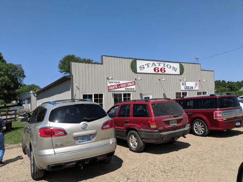 A roadside market named "Station 66" with signs for local fruits, vegetables, and ice cream, surrounded by parked cars.