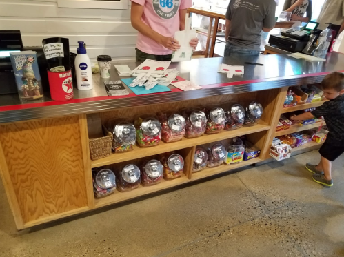 A wooden counter with gumball machines and snacks, a child reaching for candy, and a person at the register.