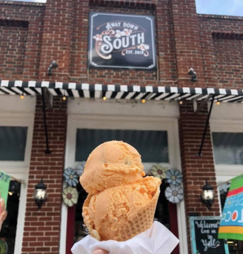 A hand holds a double scoop of orange ice cream in a cone, with a brick building and sign in the background.