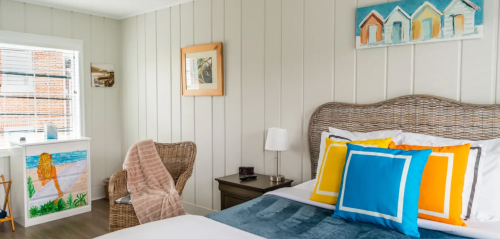 Cozy bedroom with a woven headboard, colorful pillows, and beach-themed decor, featuring a chair and window light.