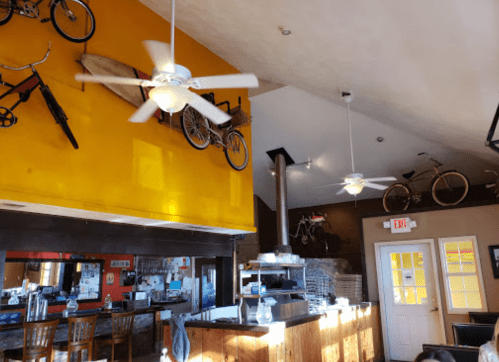 Interior of a cozy café featuring bicycles and a surfboard on the yellow wall, with a bar and ceiling fans visible.