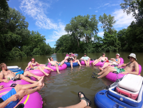 A group of people relaxing on pink inner tubes in a river, surrounded by trees and a clear blue sky.
