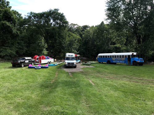 A grassy area with parked vehicles, including a blue bus, and colorful inflatable boats nearby. Trees in the background.