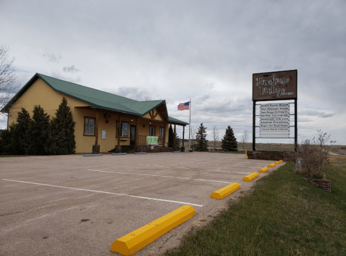 A building with a green roof and American flag, featuring a sign and parking lot, set against a cloudy sky.