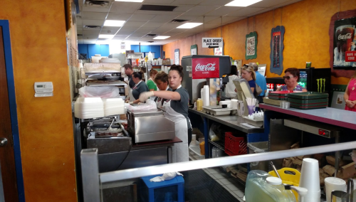 A busy restaurant kitchen with staff preparing food and customers waiting at the counter. Colorful decor in the background.