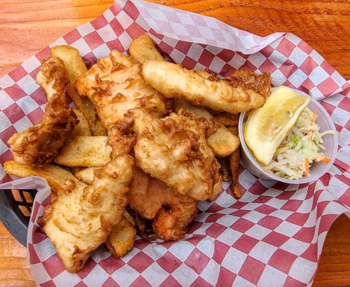 A basket of fried fish fillets with French fries, a lemon wedge, and a small cup of coleslaw on a checkered paper.