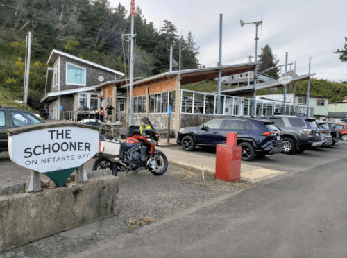 A restaurant called "The Schooner" on Netarts Bay, with parked cars and a motorcycle in front. Trees in the background.