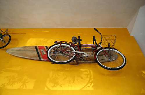 A red bicycle with a basket rests on a surfboard against a bright yellow wall.