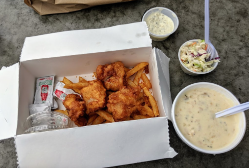 A takeout box with fried fish and fries, accompanied by coleslaw and a cup of chowder on a table.