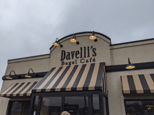 Sign for Davelli's Bagel Café on a building with striped awnings and outdoor lighting. Overcast sky in the background.