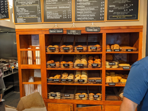 A wooden display case filled with various bagels and baked goods, with a menu board above listing options.