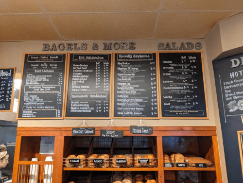 Menu board displaying bagels, sandwiches, salads, and prices at a café. Fresh baked goods visible below.