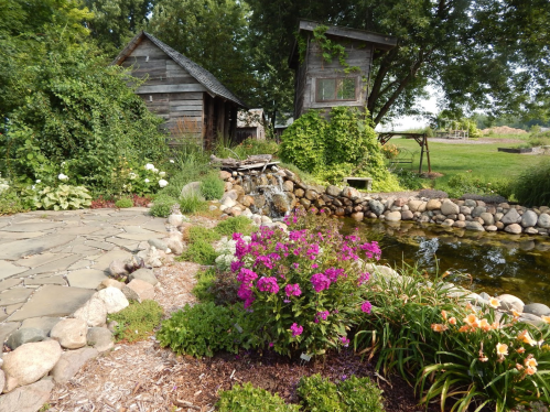 A serene garden scene featuring a pond, stone path, vibrant flowers, and rustic wooden structures surrounded by greenery.