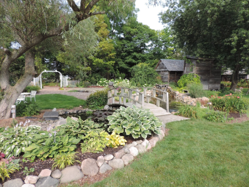 A serene garden with a wooden bridge over a pond, surrounded by lush greenery and a rustic shed in the background.