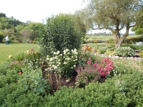 A lush garden featuring colorful flowers, greenery, and a tree, with a person walking in the background.