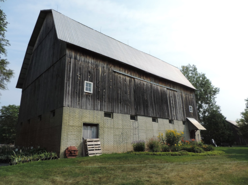 A large, weathered barn with a metal roof, surrounded by greenery and flowers on a sunny day.