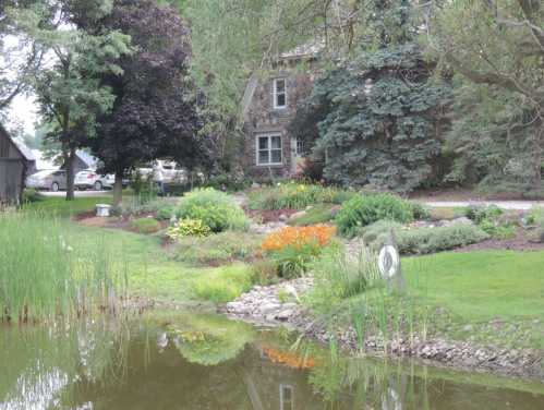 A serene garden with colorful flowers and lush greenery beside a pond, featuring a stone house in the background.