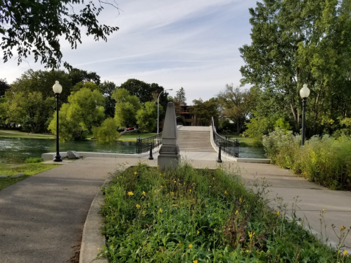 A scenic park path leads to a bridge over a calm river, surrounded by trees and greenery under a clear sky.