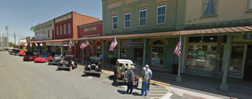 A street lined with vintage cars and shops, featuring people chatting and American flags displayed.