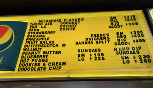 Menu board displaying milkshake flavors and sundae options with prices, including items like strawberry, banana, and peanut butter.