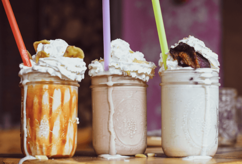 Three colorful milkshakes in jars topped with whipped cream and garnishes, featuring orange, chocolate, and vanilla flavors.