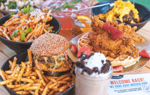A vibrant spread of various dishes, including fried chicken, burgers, loaded fries, and desserts, on a wooden table.