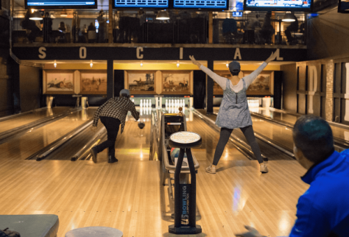 Two people bowling in a lively alley, one celebrating a strike while the other prepares to roll the ball.