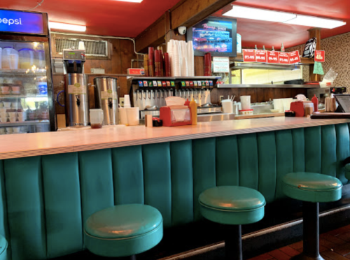 A retro diner interior featuring a turquoise counter and stools, with a drink station and menu in the background.