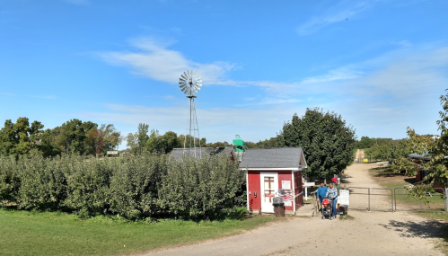 A small red building near a windmill, surrounded by trees and a dirt path, with people walking by.