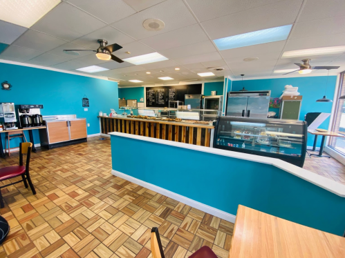 Bright café interior with blue walls, wooden counter, display case, and tiled floor, featuring tables and chairs.