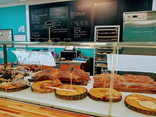A bakery display case with various breads, cakes, cookies, and macarons, featuring handwritten menu items on a chalkboard.
