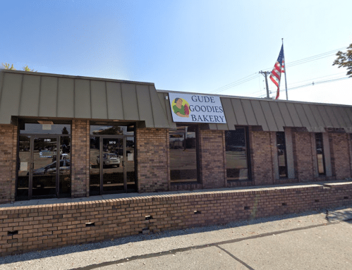Exterior of Gude Goodies Bakery, featuring a sign and an American flag, with a brick facade and large windows.
