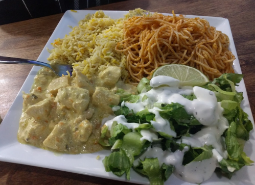 A plate with yellow rice, spicy noodles, creamy chicken curry, and a fresh salad drizzled with dressing and a lime wedge.