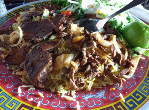 A colorful plate of seasoned rice topped with grilled meat, onions, and fresh vegetables, garnished with lime.
