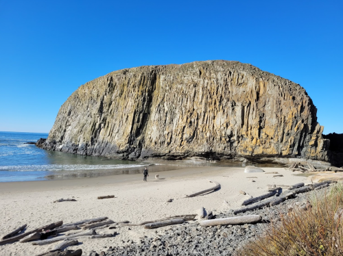 A large rocky cliff rises above a sandy beach, with a person standing near the water's edge under a clear blue sky.