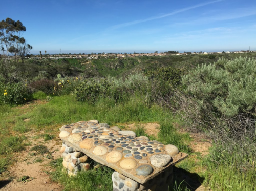 A stone bench overlooks a lush green landscape with hills and a distant view of houses and the ocean under a blue sky.