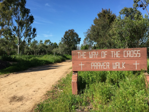 A dirt path lined with greenery, featuring a wooden sign that reads "The Way of the Cross Prayer Walk."