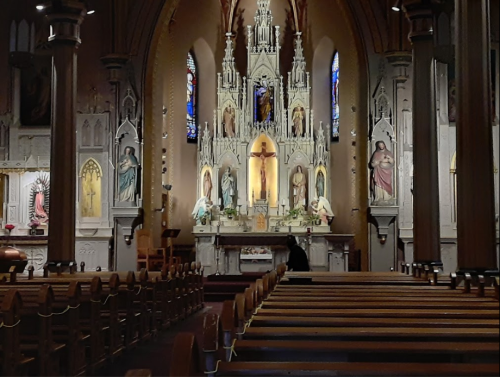 Interior of a church featuring ornate altars, stained glass windows, and wooden pews, with a figure in the background.