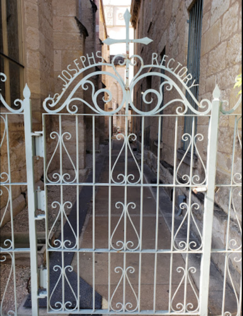 A decorative metal gate with "Joseph's Rectory" inscribed, leading to a narrow stone pathway.