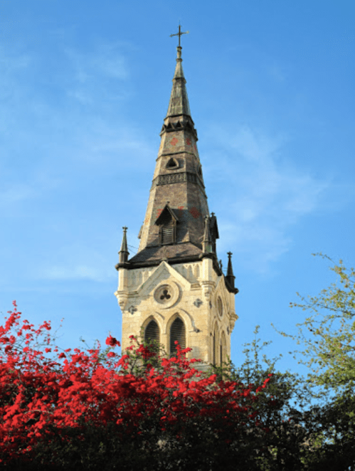 A tall church steeple rises against a blue sky, surrounded by vibrant red flowers.