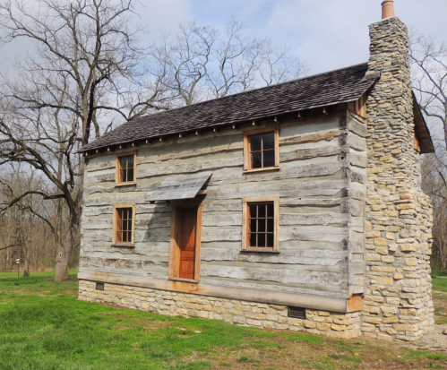 A rustic log cabin with a stone chimney, surrounded by trees and grass under a cloudy sky.