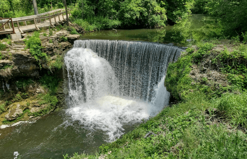 A serene waterfall cascading into a calm green pool, surrounded by lush greenery and a wooden walkway nearby.