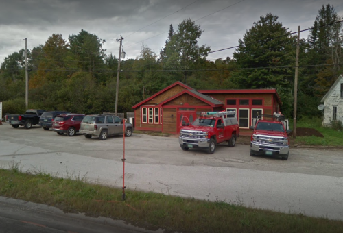 A red building with large windows, surrounded by trees, and several parked vehicles in front on a cloudy day.