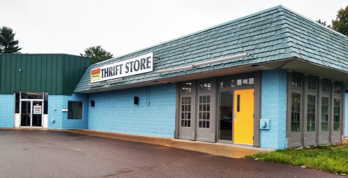 A blue thrift store building with a green roof and a bright yellow door, featuring large windows and a sign above.