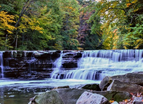 A serene waterfall cascades over rocks, surrounded by vibrant autumn foliage and a calm river.