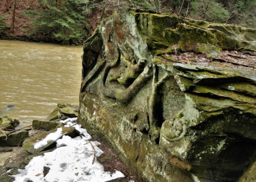 A moss-covered rock by a river, featuring intricate carvings and surrounded by trees and snow.