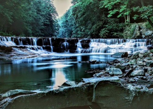 A serene waterfall cascades over rocks, surrounded by lush green trees and a calm, reflective pool of water.
