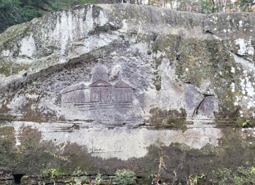 Carved stone featuring a building design, surrounded by moss and vegetation in a natural setting.
