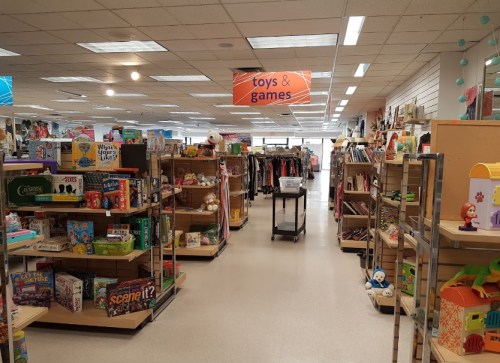 A brightly lit store aisle filled with shelves of toys, games, and clothing, with a sign reading "toys & games" above.